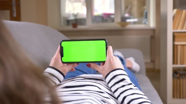 Крупним планом задній знімок жінки за допомогою телефону з зеленим екраном, що тримає його горизонтально лежачи на дивані в приміщенні в затишній квартирі — стокове відео
