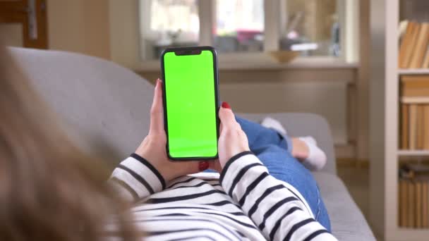 Крупный план вид сзади съемка женщины с помощью телефона с зеленым экраном держа его вертикально лежа на диване в помещении в уютной квартире — стоковое видео