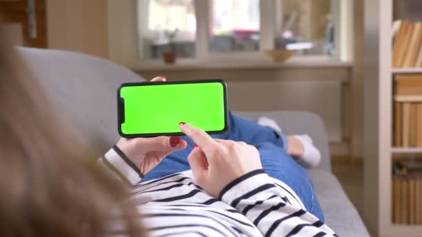 Gros plan vue arrière tournage de la mise en réseau féminine sur le téléphone avec écran vert le tenant horizontalement couché sur le canapé à l'intérieur dans un appartement confortable — Video