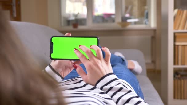 Κοντινό πλάνο γυρίσματα της γυναικείας περιήγησης στο τηλέφωνο με πράσινη οθόνη κρατώντας το οριζόντια τοποθέτηση στον καναπέ σε εσωτερικούς χώρους σε ένα φιλόξενο διαμέρισμα — Αρχείο Βίντεο