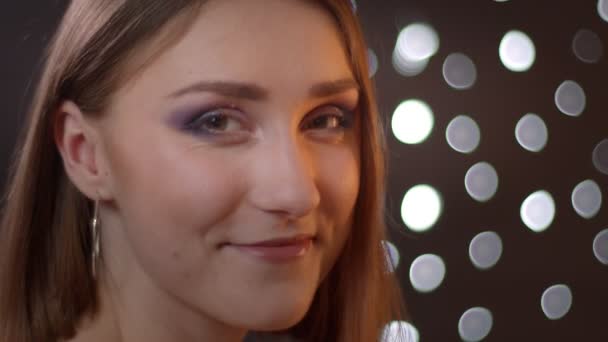 Närbild sida porträtt av unga ganska kaukasiska flicka vrida och titta på kamera leende med bokeh lampor på bakgrunden — Stockvideo