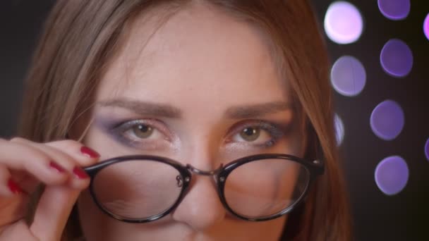 Primer plano retrato de la joven chica bastante caucásica fijación de sus gafas y levantar la ceja mirando a la cámara coqueteando felizmente con el fondo bokeh — Vídeo de stock