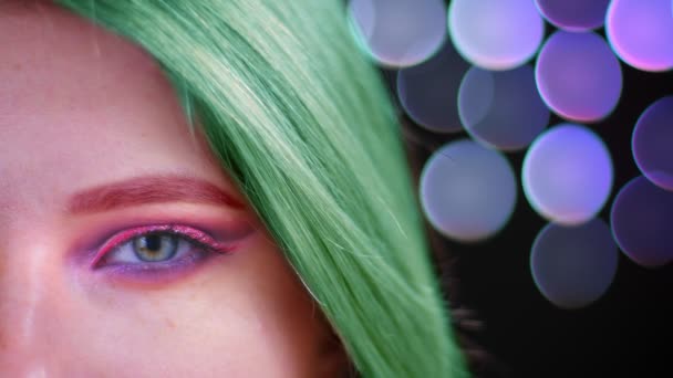 Κοντινό πλάνο μισό πρόσωπο γυρίσματα της νεαρής ελκυστική Καυκάσιος θηλυκό με ροζ όμορφο ροζ μακιγιάζ λάμψη με πράσινα βαμμένα μαλλιά κοιτάζοντας την κάμερα με bokeh φόντο — Αρχείο Βίντεο