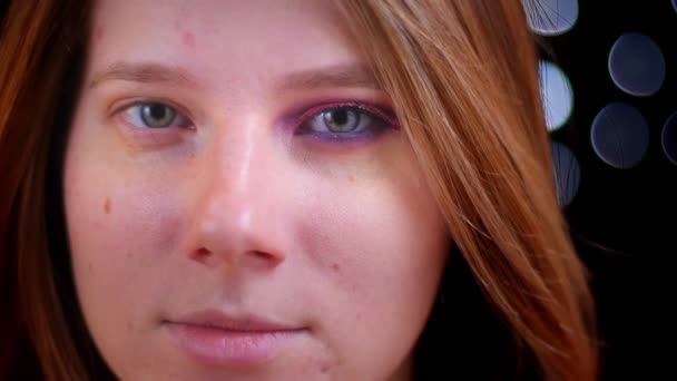Primer plano de brote de joven atractiva hembra descubriendo maquillaje medio aplicado mirando directamente a la cámara con fondo bokeh — Vídeo de stock
