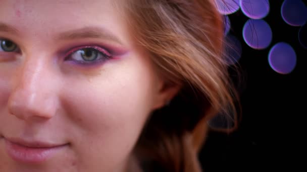 Primer plano de brote de joven atractiva cara femenina caucásica con medio maquillaje de brillo aplicado mirando a la cámara con fondo bokeh — Vídeos de Stock