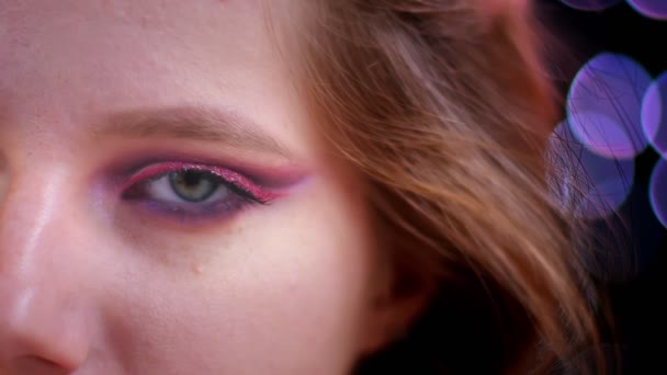 Close-up shoot van jonge aantrekkelijke Kaukasische vrouwelijke gezicht met half toegepast glitter roze make-up kijken naar camera met bokeh achtergrond — Stockvideo