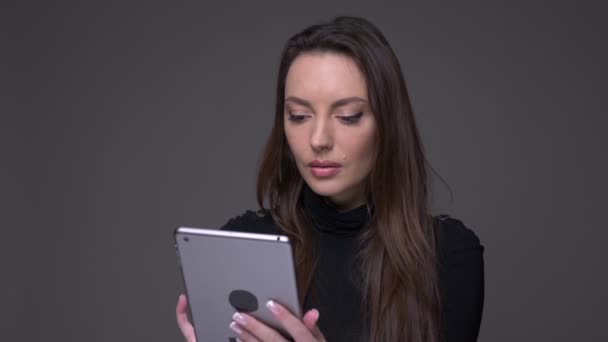 Retrato de mujer morena atractiva sosteniendo tableta mirando en la cámara en serio y fijamente aislado sobre fondo gris . — Vídeo de stock