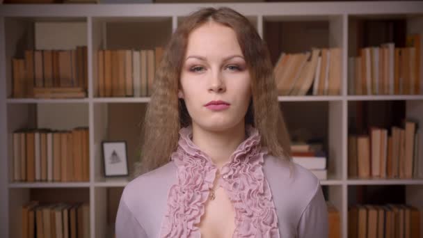 Porträt der kaukasischen welligen blonden Bibliothekarin, die ruhig und starr in die Kamera auf dem Hintergrund der Bücherregale blickt. — Stockvideo
