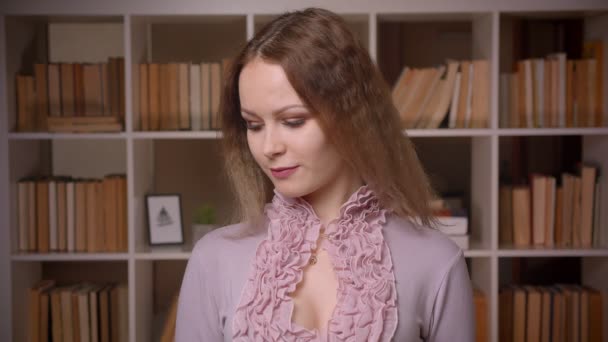 Ritratto di insegnante bionda caucasica dai capelli ondulati che posa timidamente e seducentemente nella macchina fotografica in biblioteca . — Video Stock