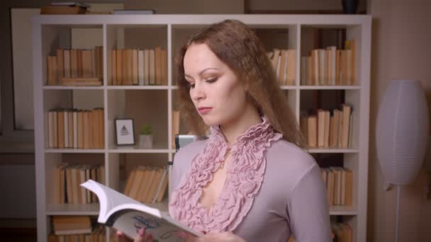 Retrato de una rubia caucásica de pelo ondulado leyendo el libro estando atenta y concentrada en la biblioteca . — Vídeo de stock