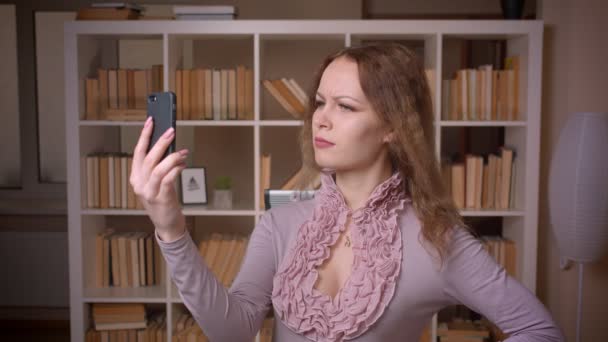 Portrét kavkazské vlnité blonďaté blond učitelky, mluvící v videochonu na smartphone s úsměvem v knihovně. — Stock video