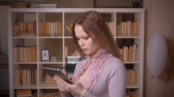 Porträtt av kaukasiska vågig blond lärare tittar på tabletten är uppmärksam och eftertänksamt på biblioteket. — Stockvideo