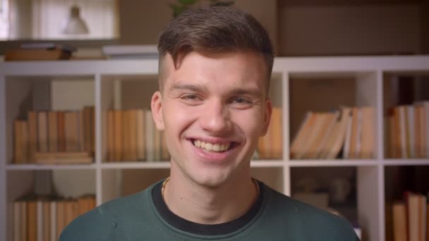 Retrato de un joven estudiante masculino sonríe en la cámara siendo extremadamente feliz en la biblioteca . — Vídeos de Stock