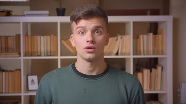 Portret van jonge mannelijke student toont sterk amusement in de camera in de bibliotheek. — Stockvideo
