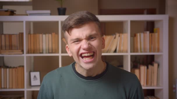 Портрет молодого студента робить смішні обличчя в камеру радісними і збудженими в бібліотеці . — стокове відео