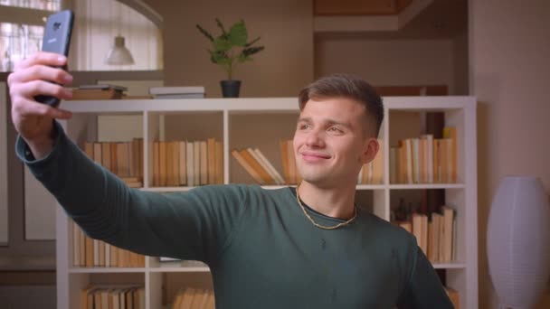 Porträtt av unga manliga studerande gör selfies på smartphone är positiv och glad på biblioteket. — Stockvideo