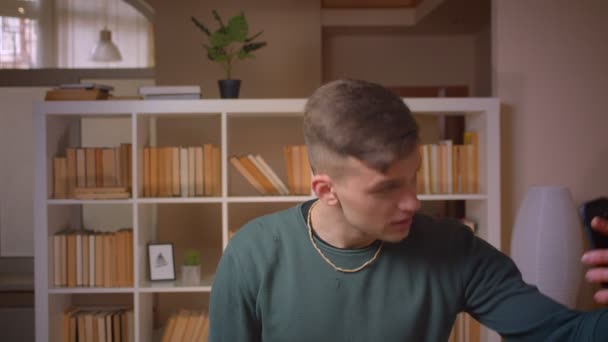 Porträtt av ung manlig student med videocall med vän på smartphone att vara glad på biblioteket. — Stockvideo