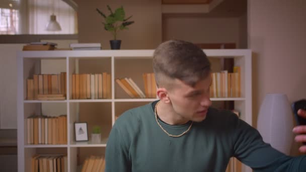 Portret młodego studenta z videocall z przyjacielem na smartfonie jest radosny w bibliotece. — Wideo stockowe