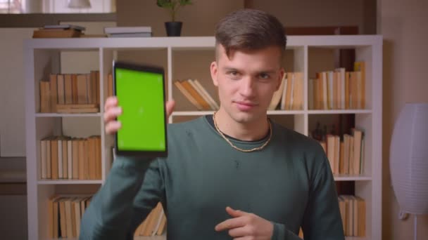 Retrato de un joven estudiante masculino enciende la aplicación en la tableta muestra la pantalla de croma verde de la misma y el pulgar hacia arriba en la biblioteca . — Vídeos de Stock