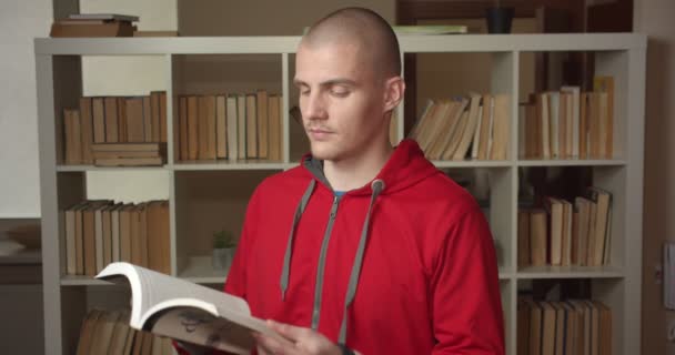 Closeup portrét mladého atraktivního bělošského studenta, který čte knihu a dívá se na kameru a usmívá se v univerzitní knihovně uvnitř — Stock video