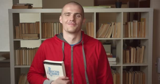Retrato de close-up do jovem estudante masculino caucasiano atraente segurando um livro olhando para a câmera e sorrindo na biblioteca da faculdade dentro de casa — Vídeo de Stock