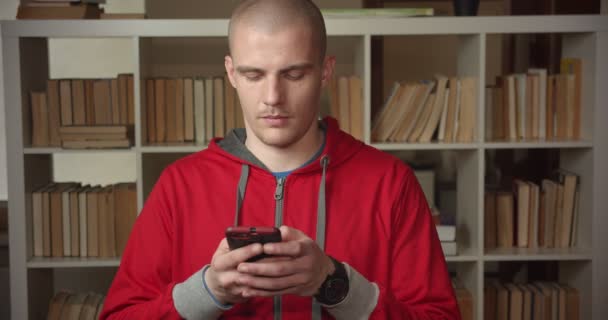 Nahaufnahme Porträt einer jungen attraktiven kaukasischen männlichen Studenten mit dem Telefon in der Universitätsbibliothek drinnen — Stockvideo