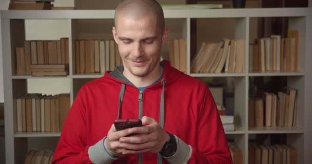 Retrato de close-up do jovem estudante masculino caucasiano atraente usando o telefone olhando para a câmera e sorrindo na biblioteca da faculdade dentro de casa — Vídeo de Stock