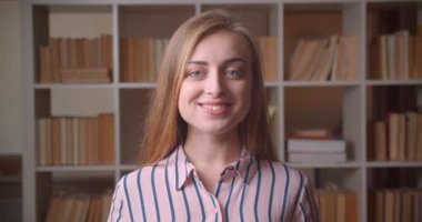 Genç güzel beyaz kadın öğrencinin fotoğraf makinasına bakıyor, üniversite kitaplığında mutlu bir şekilde gülümsüyor