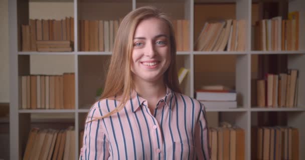 Gros plan portrait de jeune étudiante caucasienne joyeuse souriant avec excitation en regardant la caméra dans la bibliothèque du collège à l'intérieur — Video