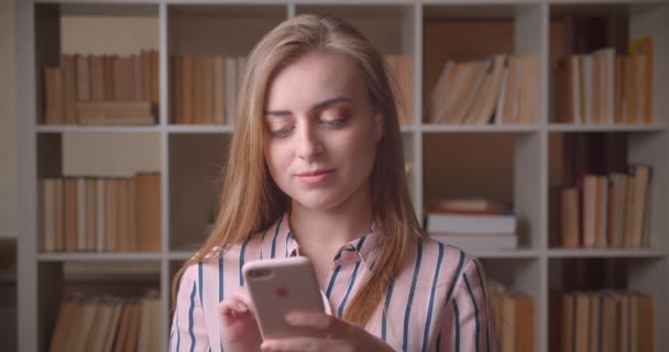 Closeup portrait of young pretty caucasian female student using the phone in the college library — Stock Video