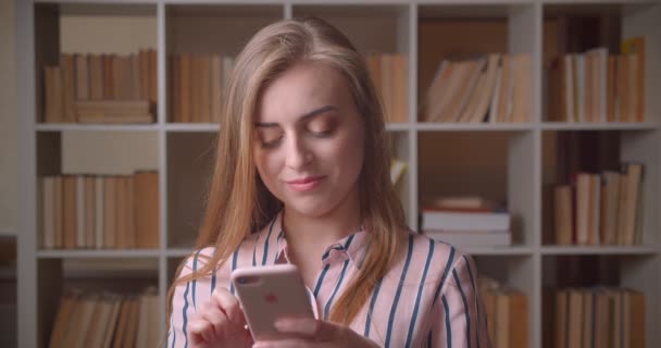 Κοντινό πλάνο του νεαρού όμορφο καυκάσιο θηλυκό μαθητή μηνύματα στο τηλέφωνο στη βιβλιοθήκη κολλέγιο — Αρχείο Βίντεο