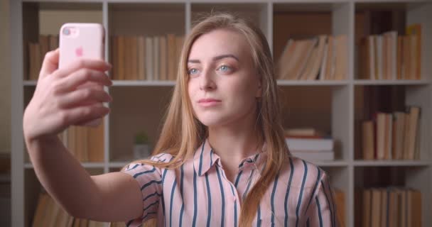 Primo piano ritratto di giovane studentessa caucasica che si fa dei selfie al telefono nella biblioteca del college — Video Stock