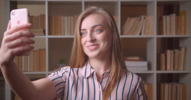 Närbild porträtt av unga vackra kaukasiska kvinnlig elev som har ett video samtal på telefonen i College biblioteket — Stockvideo
