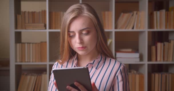 Retrato de cerca de una joven estudiante bastante caucásica usando la tableta mirando la cámara en la biblioteca de la universidad — Vídeos de Stock