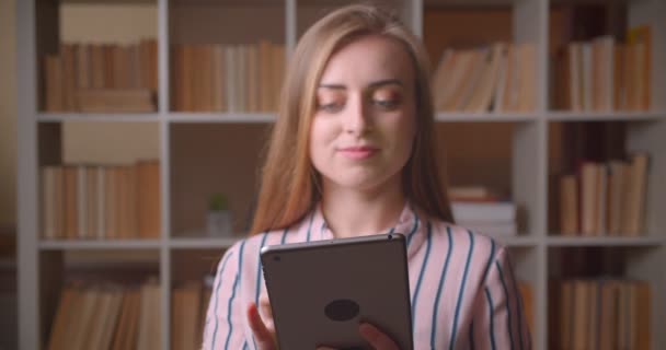 Крупный план портрета молодой симпатичной белой студентки, использующей планшет и показывающей зеленый хроматический экран на камеру в библиотеке колледжа — стоковое видео