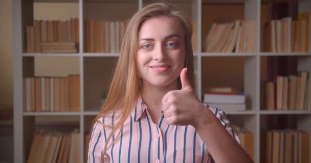 Крупный план портрета молодой симпатичной белой студентки, показывающей большой палец, улыбающейся, глядя в камеру в библиотеке колледжа — стоковое видео