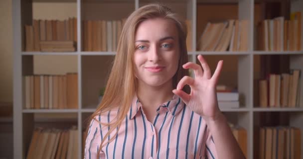 Closeup portrait of young pretty caucasian female student showing okay sign smiling looking at camera in the college library — Stock Video