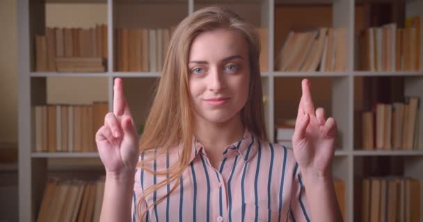Closeup portrait of young pretty caucasian female student having her fingers crossed being hopeful looking at camera in the college library — Stock Video