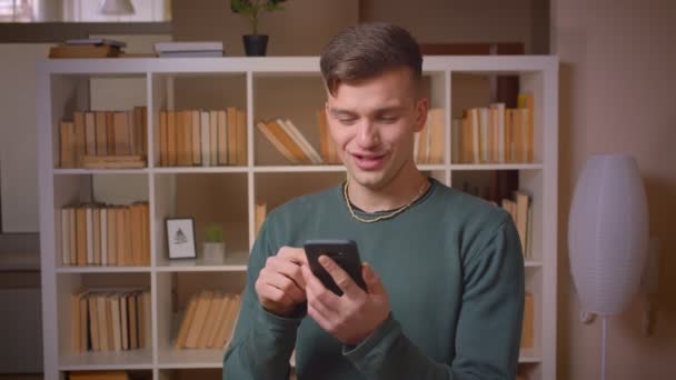 Portret młodego studenta płci męskiej oglądając uważnie w smartfonie jest uprzejmy i emocjonalny w bibliotece. — Wideo stockowe