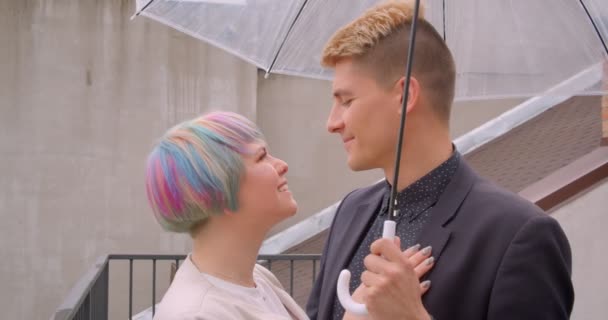 젊은 아름다운 힙스터 커플의 클로즈업 촬영은 행복한 야외에서 우산 아래에 함께 서있는 서로를 보고 — 비디오