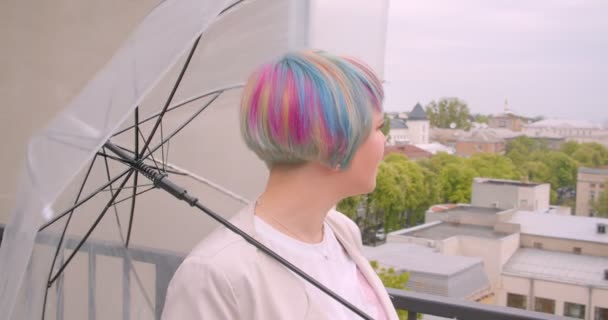 Nahaufnahme Porträt einer jungen hübschen kaukasischen Frau mit gefärbten Haaren, die einen Regenschirm hält und auf ihren Freund im Freien mit landschaftlichem Hintergrund wartet — Stockvideo
