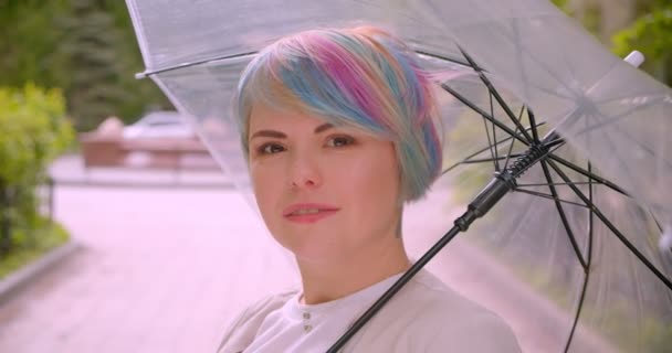 公園で楽しそうに笑顔のカメラを見て傘を持った髪を染めた若いかわいい白人女性のクローズアップポートレート — ストック動画