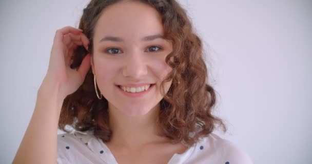 Κοντινό γύρισμα της νεαρής όμορφη σγουρά Καυκάσιος θηλυκό χαμογελάει ευχαρίστως φτιάχνοντας τα μαλλιά της κοιτάζοντας την κάμερα με φόντο απομονώνεται σε λευκό — Αρχείο Βίντεο