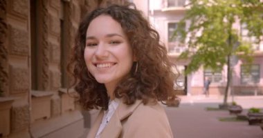 Sokakta açık havada duran kameraya mutlu bir şekilde bakan genç uzun saçlı kıvırcık beyaz beyaz kız öğrencinin closeup portresi