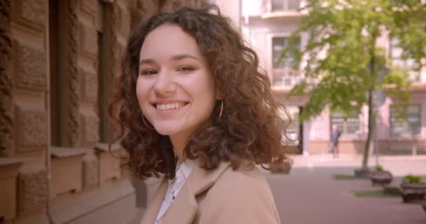 Nahaufnahme Porträt der jungen langhaarigen lockigen kaukasischen Studentin, die glücklich in die Kamera lächelt, die draußen auf der Straße steht — Stockvideo
