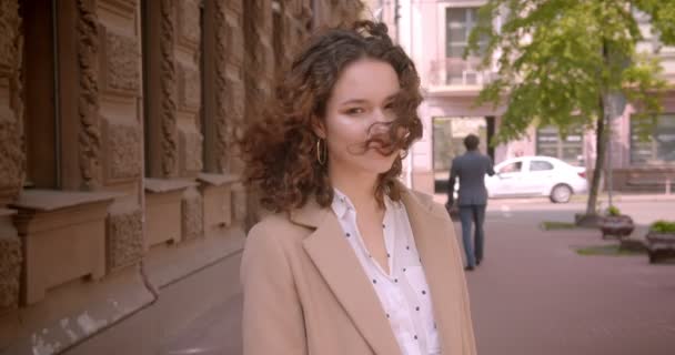 Gros plan portrait de jeune étudiante caucasienne bouclée aux cheveux longs souriant posant joyeusement devant la caméra debout à l'extérieur dans la rue de la ville — Video