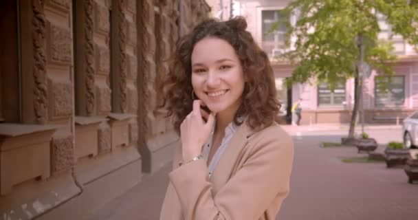 Крупный план портрета молодой кудрявой белокурой студентки, счастливо улыбающейся перед камерой, стоящей на улице в городе — стоковое видео