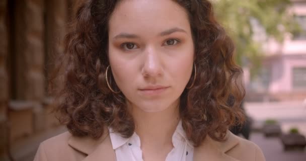 Close-up shoot van jonge langharige krullend blanke vrouwelijke student kijkend naar camera staand buitenshuis op straat — Stockvideo