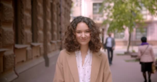 Närbild shoot av unga långhåriga lockig kaukasiska kvinnlig student leende glatt gå mot kameran utomhus på gatan med suddig fokus — Stockvideo