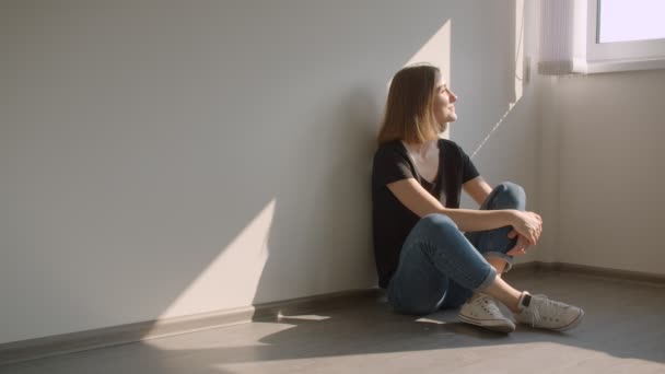 Крупный план портрета молодой симпатичной белой женщины, сидящей на полу у окна и счастливо улыбающейся в уютной солнечной квартире — стоковое видео